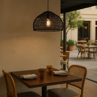 Black Frya 40 cm hand-woven rattan pendant light hanging above a wooden dining table in a stylish restaurant interior with open-air seating in the background, featuring natural textures, timber furniture, and warm ambient lighting in a relaxed coastal setting.