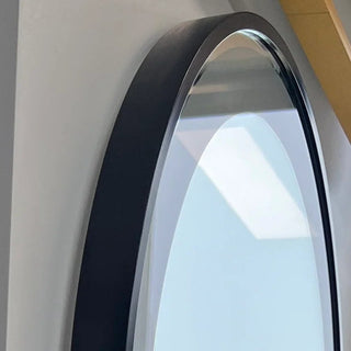 A tall, arched mirror with a black frame mounted on a wall inside a vehicle or building, reflecting an outdoor scene with a vehicle and parking lot.