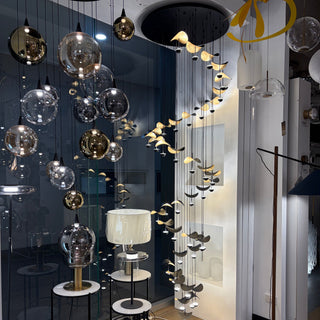 A modern interior showroom featuring a large decorative hanging light fixture with many clear glass spheres and small bird-shaped bulbs. The room has wood flooring, white walls, and additional lamps and hanging lights visible in the background.