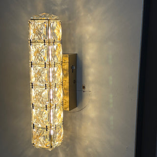 A modern cylindrical wall sconce with a textured, warm white glass body and a brushed gold metal frame, mounted on a light gray wall and casting a soft glow.