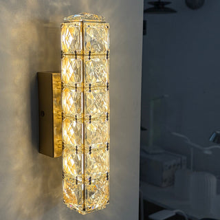 A modern cylindrical wall sconce with a warm glowing crystal prismatic column and a brushed gold metal base, mounted on a light-colored wall.