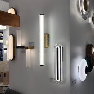 A display of modern wall sconces mounted on a light gray wall in a showroom. Visible designs include a tall vertical bar with a frosted glass shade, a slim rectangular light with black and white horizontal bars, a gold-finished vertical bar, and several other contemporary styles Displayed at Davoluce Lighting showroom in Elsternwick
