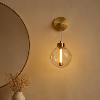 Wall-mounted light fixture with a clear glass shade on a beige wall, next to a round mirror and small table with decor items.