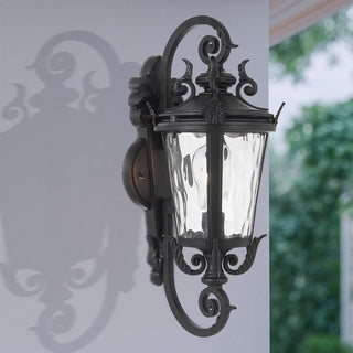 Telbix PEDRAL Decorative outdoor wall light fixture on a white wall with blurred greenery in the background