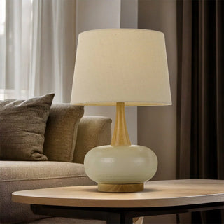 Table lamp with beige shade and green base on a wooden table, with a sofa and curtains in the background.