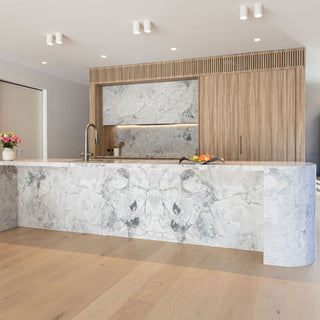 A modern kitchen island featuring a white and gray marble countertop and matching marble backsplash, set in a bright room with light wood flooring and slatted wood cabinetry.