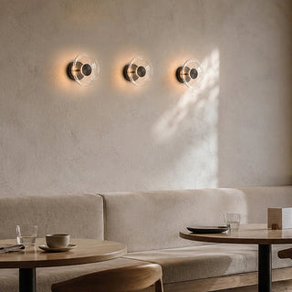Three modern wall sconces with clear circular glass shades and dark metal centers are mounted on a textured beige wall above a light upholstered bench and wooden tables in a softly lit interior.