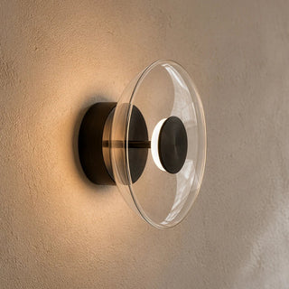 A modern wall-mounted light fixture with a clear glass dome and a matte black metal base, shown lit against a textured beige wall.